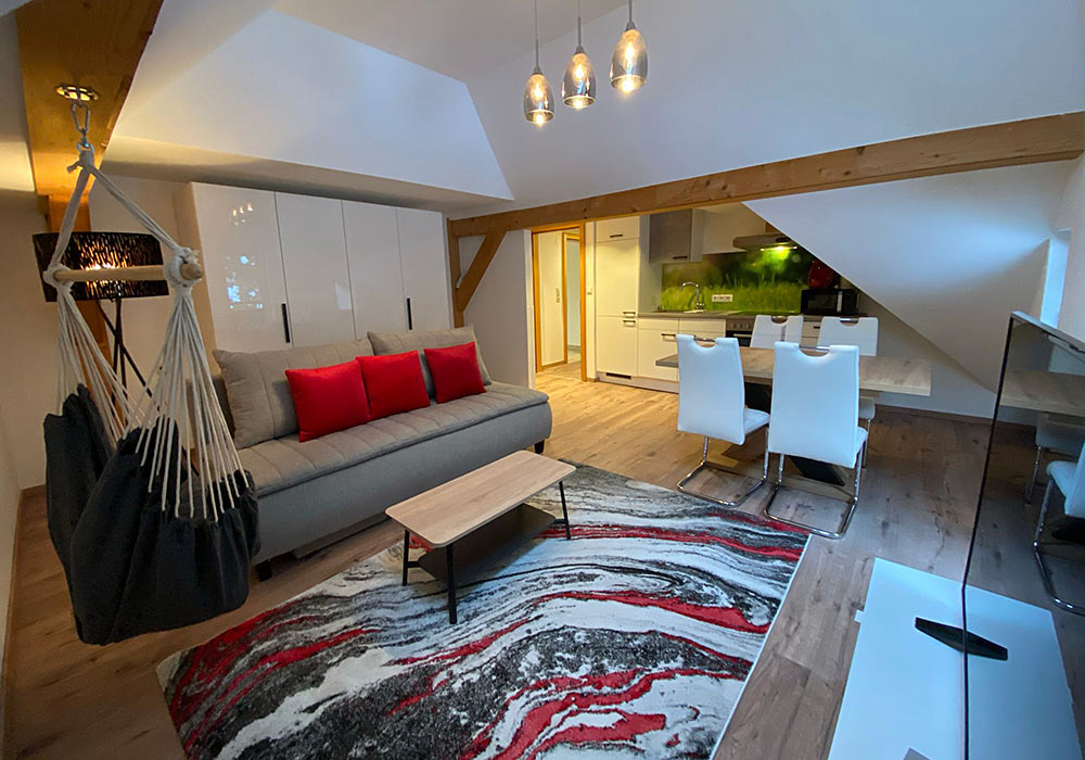 ankogel_Ferienwohnung_GRUBERS_Hotel_Apartments_Gastein Appartement Ankogel Wohnzimmer mit Hängesessel, Teppich rot-weiß-grau, grauem Sofa und Essecke mit weißen Stühlen