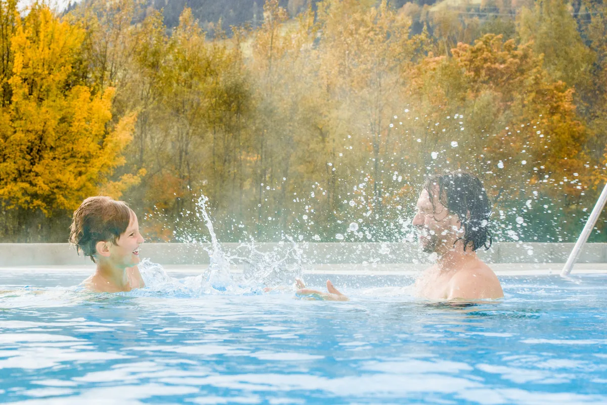 Vater und Kind spielen im Thermalwasser vor goldenen Herbstbäumen in Gastein