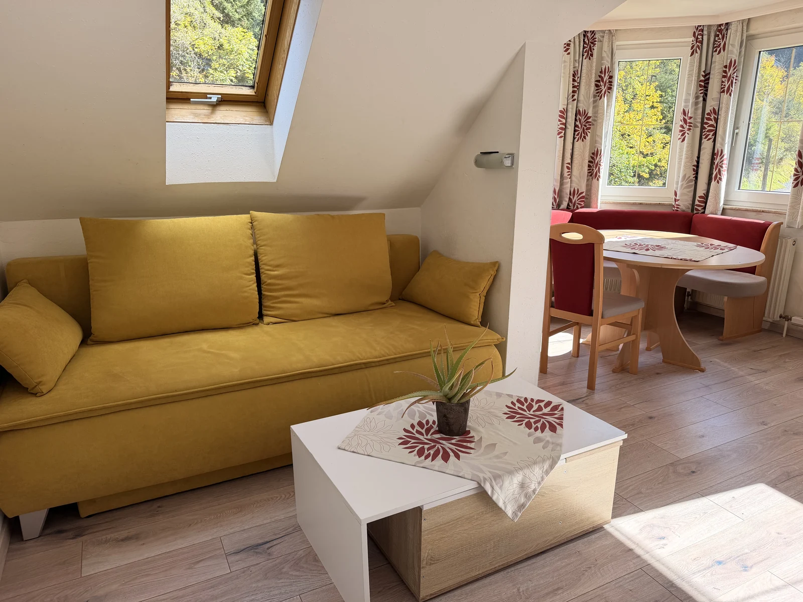 Helles Wohnzimmer der Ferienwohnung Graukogel mit gelbem Sofa, Couchtisch und Essecke am Fenster