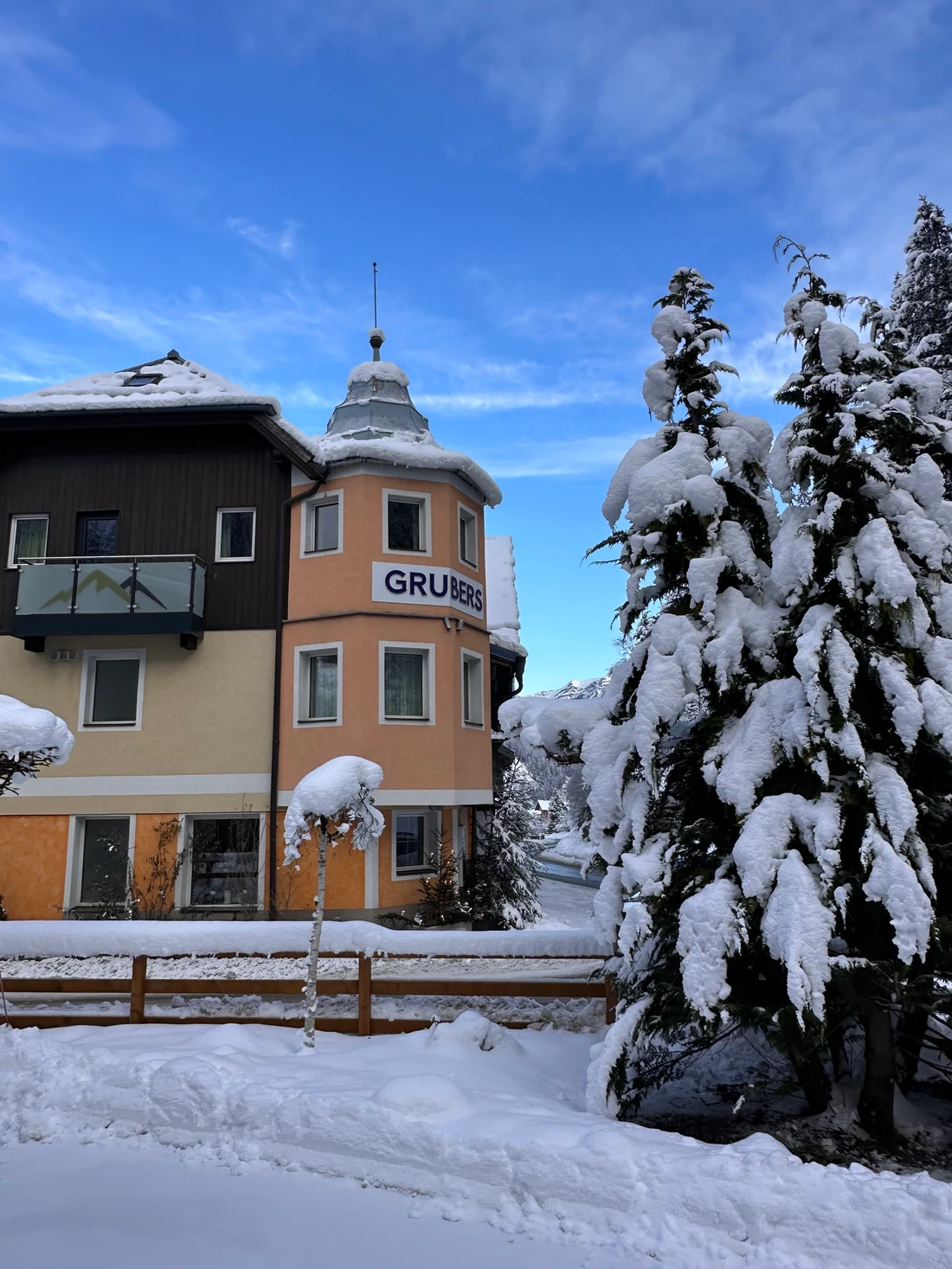 GRUBERS in Bad Gastein-Böckstein im Winter, mit Erker, Balkon und schneebedecktem Baum vor blauem Himmel.